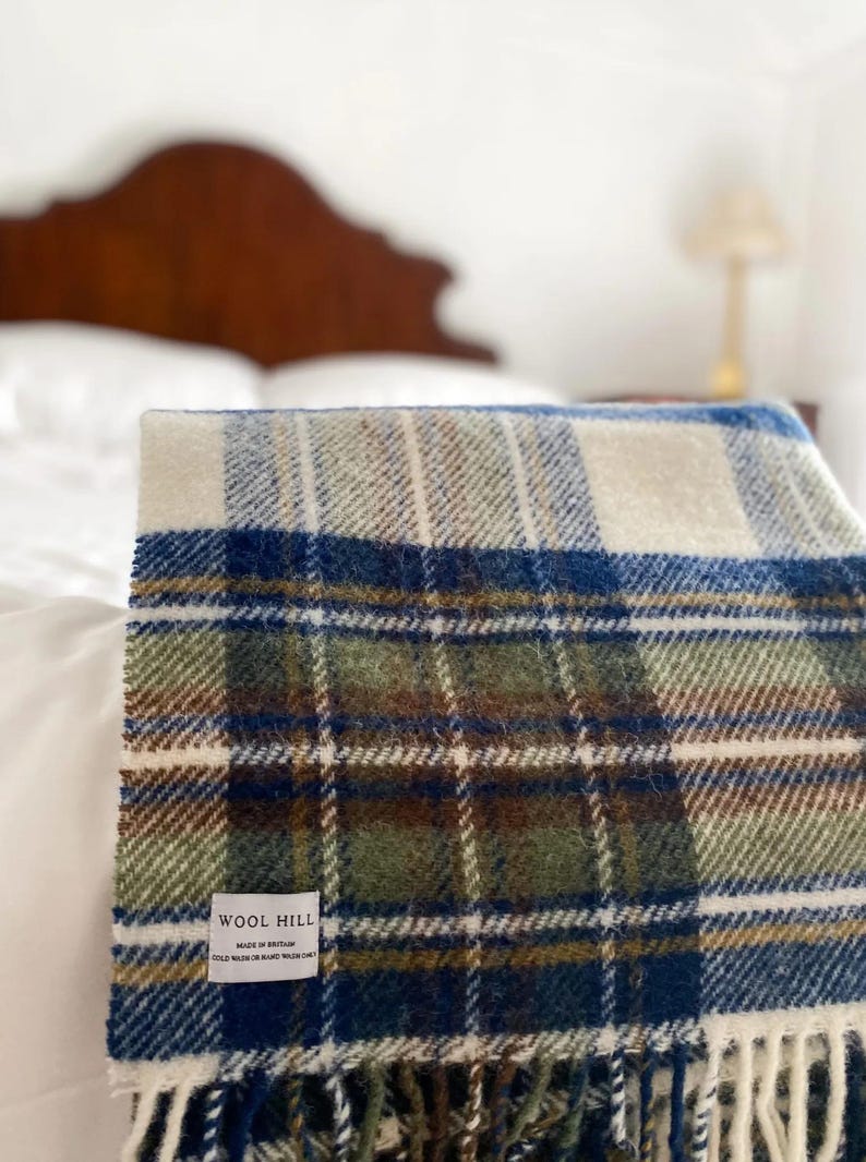 Puede incluir: Una manta de lana a cuadros blancos, azules, verdes y marrones con flecos. La manta tiene una etiqueta que dice "Wool Hill Made in Britain 100% Wool Hand Wash Only".