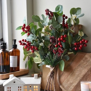 May include: A festive bouquet of faux greenery with red berries, pinecones, and sprigs, tied with twine. The arrangement sits on a marble surface near white ceramic house-shaped candle holders and amber glass soap dispensers. A wooden cutting board is in the background.