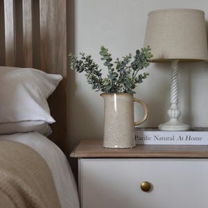 May include: A white wooden bed frame with a white pillow and a beige blanket. A white nightstand with two drawers and gold knobs sits next to the bed. A ceramic pitcher with a speckled glaze holds a bouquet of green eucalyptus branches. A lamp with a beige linen shade sits on the nightstand. The book "Pacific Natural At Home" is on the nightstand.