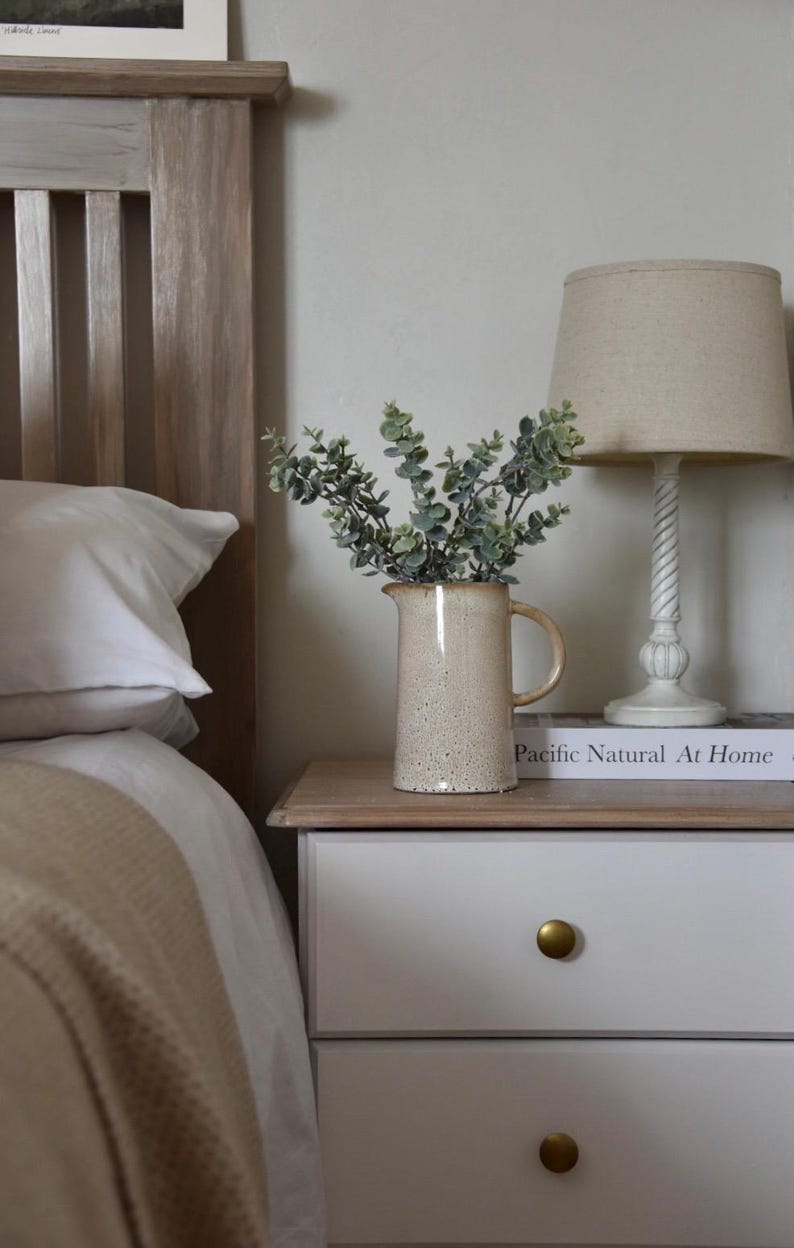 May include: A white wooden bed frame with a white pillow and a beige blanket. A white nightstand with two drawers and gold knobs sits next to the bed. A ceramic pitcher with a speckled glaze holds a bouquet of green eucalyptus branches. A lamp with a beige linen shade sits on the nightstand. The book "Pacific Natural At Home" is on the nightstand.