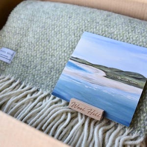 K&ouml;nnte beinhalten: Eine gr&uuml;ne und wei&szlig;e Wolldecke mit Fransenbord&uuml;re, gefaltet in einem Karton. Auf der Decke liegt eine kleine Karte mit einem Gem&auml;lde einer Strandszene. Die Karte tr&auml;gt den Text "Wool Hill".