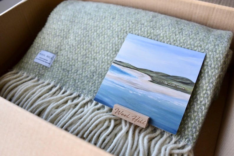 K&ouml;nnte beinhalten: Eine gr&uuml;ne und wei&szlig;e Wolldecke mit Fransenbord&uuml;re, gefaltet in einem Karton. Auf der Decke liegt eine kleine Karte mit einem Gem&auml;lde einer Strandszene. Die Karte tr&auml;gt den Text "Wool Hill".