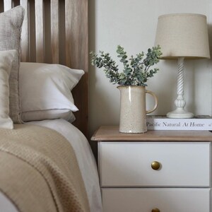 May include: A white nightstand with three drawers and gold knobs. A beige ceramic pitcher with a bouquet of green eucalyptus sits on top of the nightstand, next to a book titled "Pacific Natural At Home".