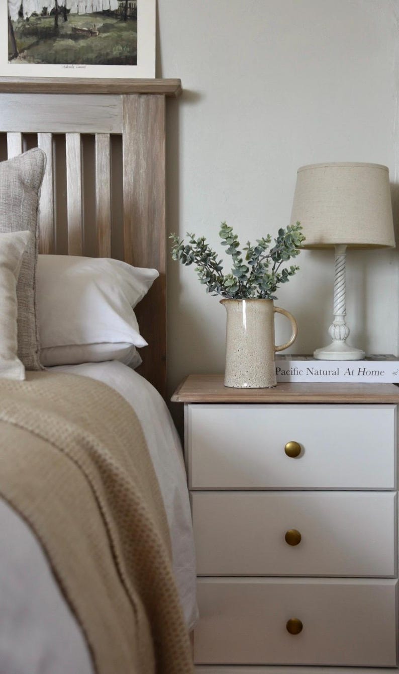 May include: A white nightstand with three drawers and gold knobs. A beige ceramic pitcher with a bouquet of green eucalyptus sits on top of the nightstand, next to a book titled "Pacific Natural At Home".