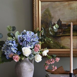 Pu&ograve; includere: Una composizione floreale in un vaso grigio con ortensie blu, rose bianche e bacche rosa. Un dipinto incorniciato e una candela alta sono sullo sfondo. Libri e una forma di scarpa in legno sono su una superficie di legno.