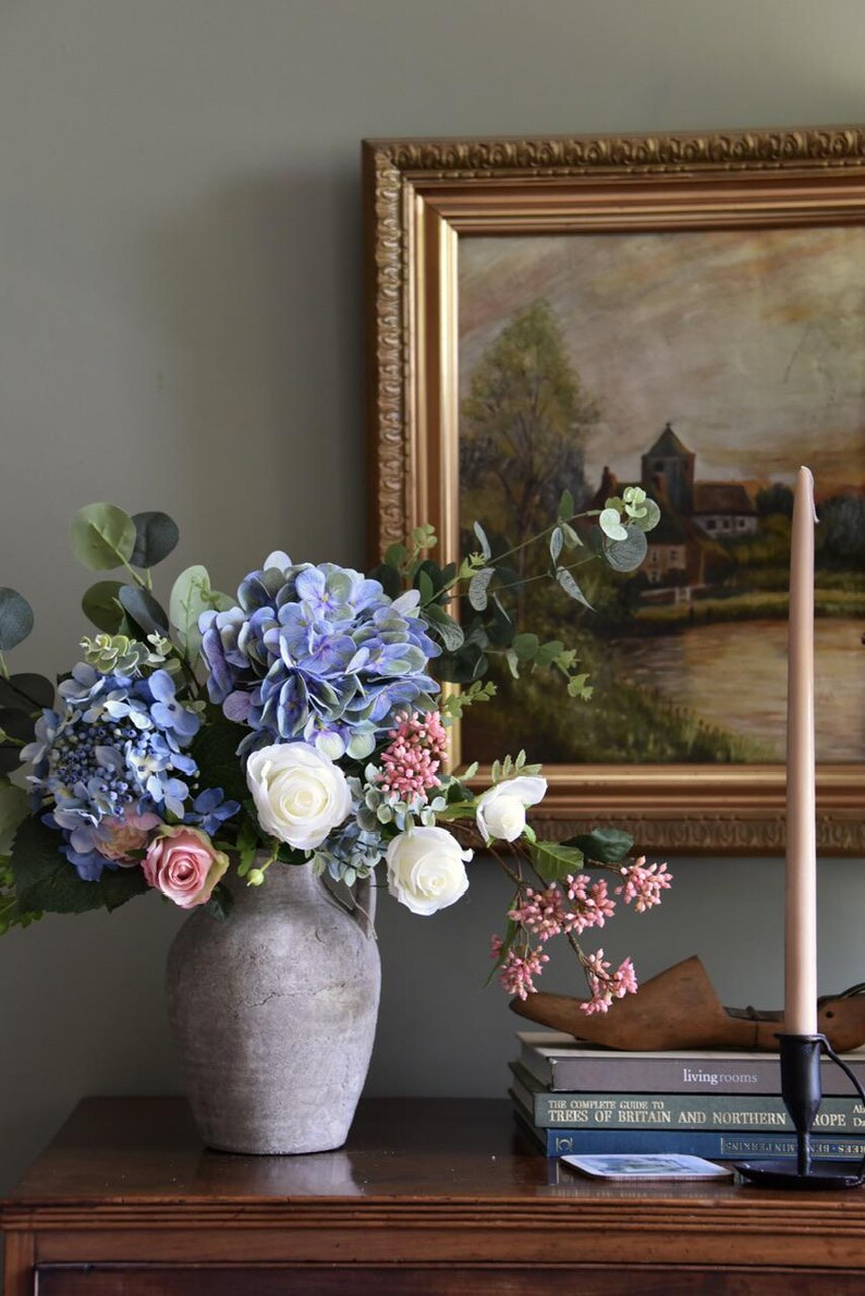 Pu&ograve; includere: Una composizione floreale in un vaso grigio con ortensie blu, rose bianche e bacche rosa. Un dipinto incorniciato e una candela alta sono sullo sfondo. Libri e una forma di scarpa in legno sono su una superficie di legno.