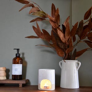 May include: A white ceramic oil diffuser with a small candle inside. The diffuser is lit and has a warm glow. The diffuser is on a wooden surface with a brown bottle of soap and a vase with dried leaves.