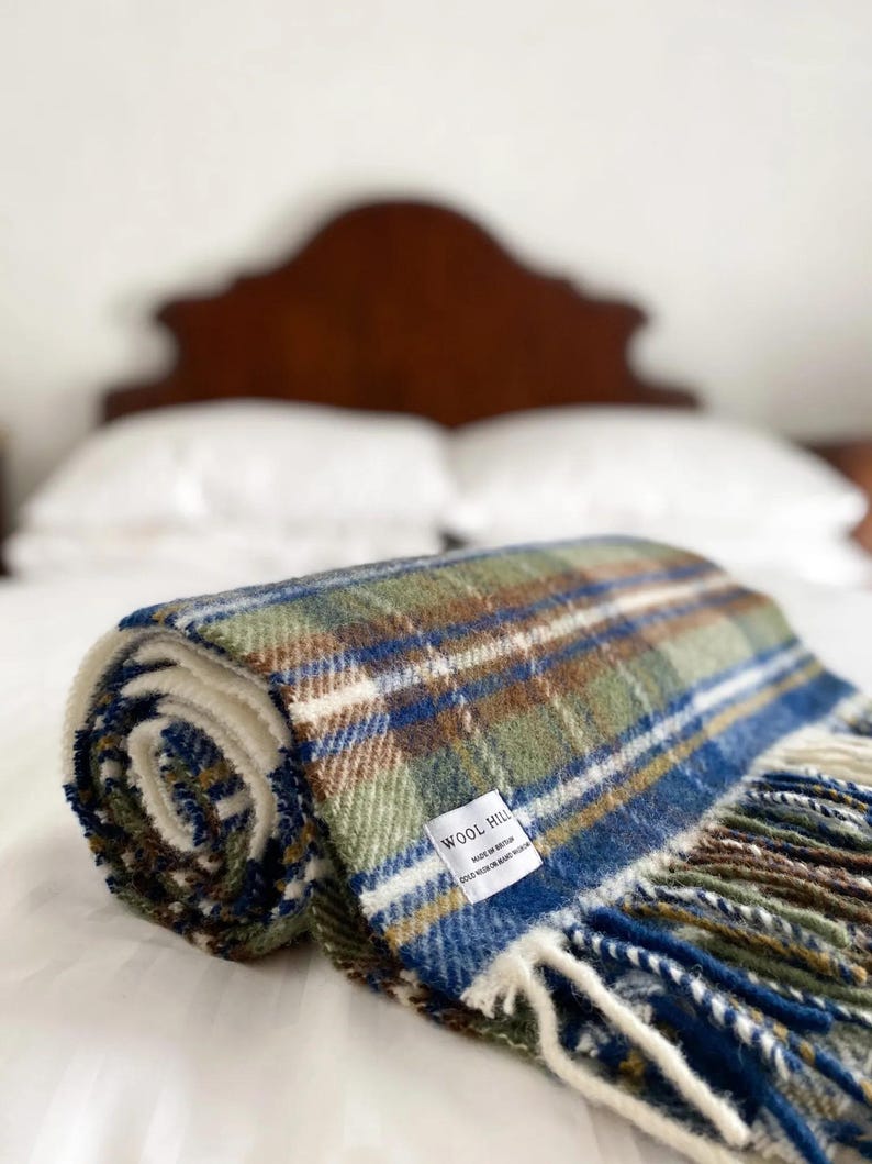Puede incluir: Una manta de lana a cuadros verde, azul y marr&oacute;n enrollada con flecos. La manta tiene una etiqueta blanca que dice "Wool Hill" y "Made in Britain 100% New Zealand Wool".
