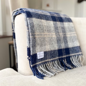 Puede incluir: Una manta de lana a cuadros azules y blancos con flecos en los bordes. La manta est&aacute; doblada y drapeada sobre un sill&oacute;n blanco. La manta tiene una peque&ntilde;a etiqueta que dice "Wool Hill Made in Britain".