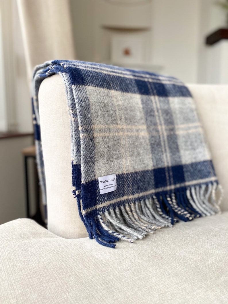 Puede incluir: Una manta de lana a cuadros azules y blancos con flecos en los bordes. La manta est&aacute; doblada y drapeada sobre un sill&oacute;n blanco. La manta tiene una peque&ntilde;a etiqueta que dice "Wool Hill Made in Britain".