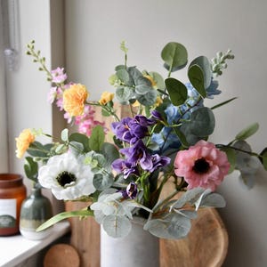 Könnte beinhalten: Ein Strauß künstlicher Blumen in einer grauen Keramikvase. Der Strauß enthält rosa, weiße, lila und blaue Blumen sowie grüne Eukalyptusblätter.