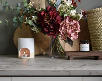 May include: A ceramic essential oil diffuser with a lit tea light, a bouquet of burgundy and pink hydrangeas in a glass vase, and a small jar labeled "Bergamot" on a wooden tray. A woven basket and wooden cutting boards are in the background.