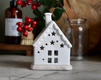 May include: A white ceramic house-shaped candle holder with star cutouts and a chimney. The house has a scalloped roofline and sits on a light-colored base. Red berries and green leaves are in the background.
