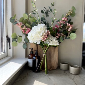 May include: A bouquet of white hydrangeas and pink flowers with green leaves arranged in a wooden vase on a grey countertop.