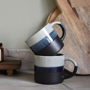 May include: Two ceramic mugs, one stacked on the other. Each mug has a black base, a blue band, and a white top. The handles are white with a blue accent. The mugs are on a marble surface.