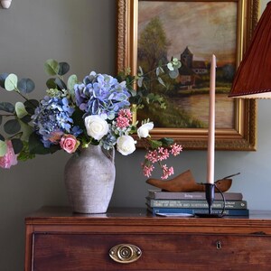 Pu&ograve; includere: Una composizione di natura morta con un bouquet di ortensie blu, rose bianche e rosa in un vaso grigio. Un dipinto incorniciato, una candela alta e libri sono su una cassettiera in legno scuro. Un paralume &egrave; visibile a destra.