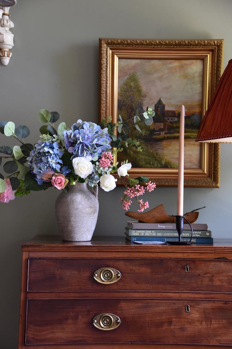 Pu&ograve; includere: Una composizione di natura morta con un bouquet di ortensie blu, rose bianche e rosa in un vaso grigio. Un dipinto incorniciato, una candela alta e libri sono su una cassettiera in legno scuro. Un paralume &egrave; visibile a destra.