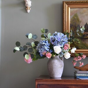 Pu&ograve; includere: Una composizione floreale in un vaso grigio con ortensie blu, rose bianche e fiori rosa, con foglie verdi di eucalipto. Il vaso &egrave; su una cassettiera in legno scuro con ferramenta in ottone.