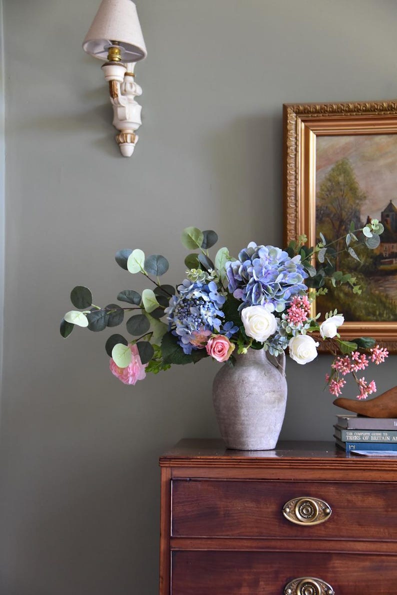 Pu&ograve; includere: Una composizione floreale in un vaso grigio con ortensie blu, rose bianche e fiori rosa, con foglie verdi di eucalipto. Il vaso &egrave; su una cassettiera in legno scuro con ferramenta in ottone.
