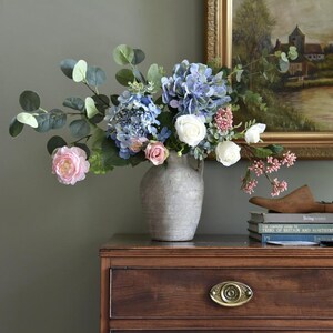 Pu&ograve; includere: Un'arrangiamento floreale in un vaso grigio su una cassettiera in legno. Il bouquet include ortensie blu, rose rosa, rose bianche e foglie di eucalipto verdi. Un dipinto incorniciato e libri sono visibili sullo sfondo.