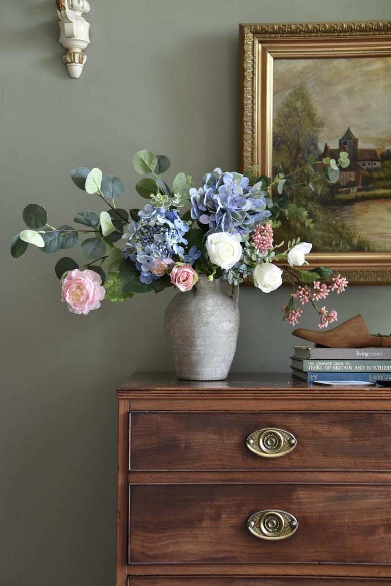Pu&ograve; includere: Un'arrangiamento floreale in un vaso grigio su una cassettiera in legno. Il bouquet include ortensie blu, rose rosa, rose bianche e foglie di eucalipto verdi. Un dipinto incorniciato e libri sono visibili sullo sfondo.