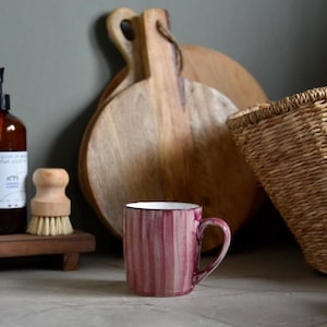 May include: A maroon and white striped ceramic mug with a curved handle. The mug is set on a light-coloured surface, with wooden chopping boards and a woven basket in the background. A wooden brush and a bottle are also visible.