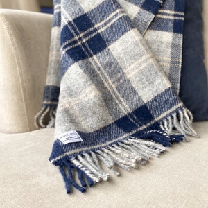 Puede incluir: Una manta de lana a cuadros azules y blancos con flecos en un sof&aacute; de color crema. La manta tiene una etiqueta que dice "Wool Hill".