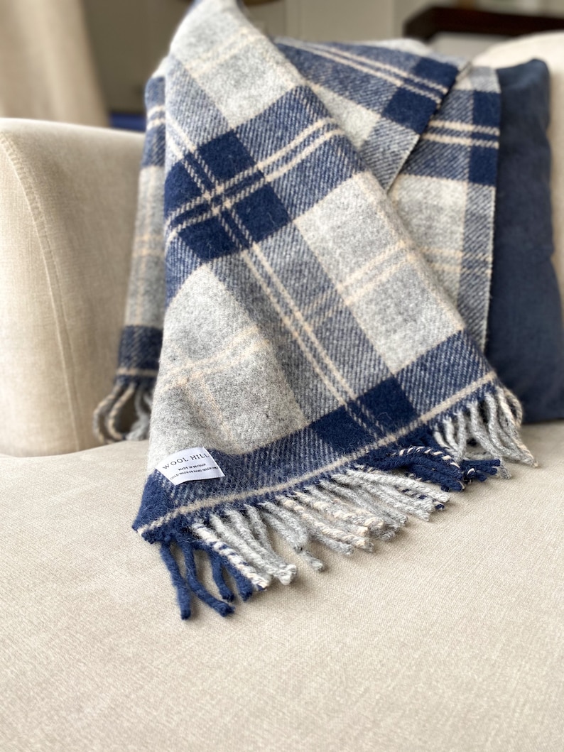 Puede incluir: Una manta de lana a cuadros azules y blancos con flecos en un sof&aacute; de color crema. La manta tiene una etiqueta que dice "Wool Hill".
