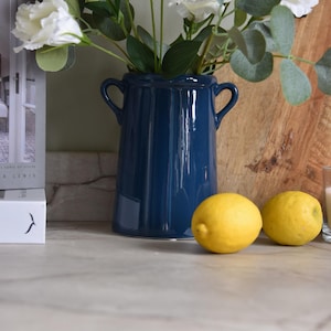 Può includere: Un vaso in ceramica blu navy lucido con due piccole maniglie laterali, contenente un bouquet di fiori bianchi e foglie di eucalipto verdi. Il vaso è posizionato accanto a due limoni gialli su una superficie chiara con venature.