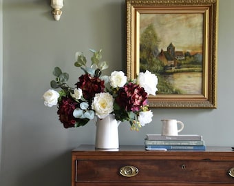 Puede incluir: Una cómoda de madera oscura con herrajes de latón sostiene una jarra blanca con flores, libros y una taza pequeña. Una pintura enmarcada cuelga encima. Se ve una lámpara de pared con una pantalla beige.