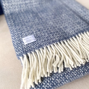 K&ouml;nnte beinhalten: Eine blaue und wei&szlig;e gewebte Wolldecke mit Fransen. Die Decke hat ein Etikett mit der Aufschrift "Wool Hill, Made in Scotland, Cold Wash or Hand Wash Only".