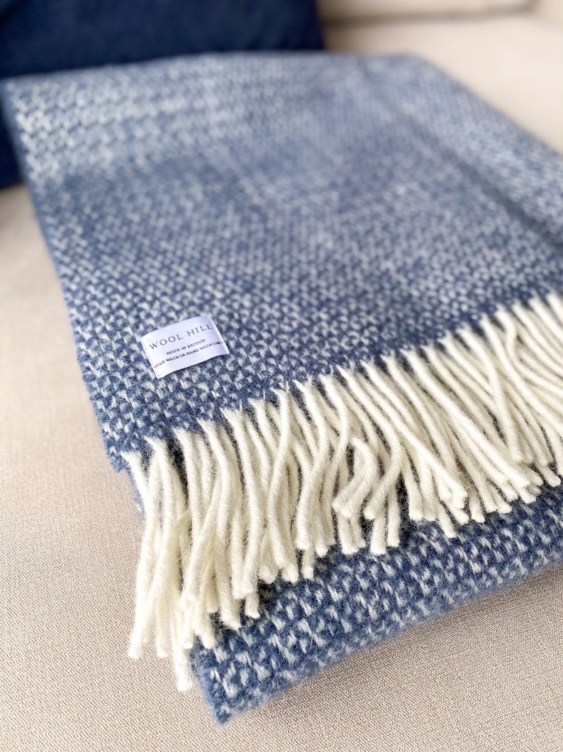K&ouml;nnte beinhalten: Eine blaue und wei&szlig;e gewebte Wolldecke mit Fransen. Die Decke hat ein Etikett mit der Aufschrift "Wool Hill, Made in Scotland, Cold Wash or Hand Wash Only".