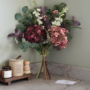 Pode incluir: Um buquê de flores artificiais com hortênsias rosa e bordô, flores brancas e folhas de eucalipto verdes. O buquê é amarrado com barbante e está sobre uma bancada de mármore com velas e uma bandeja de madeira.