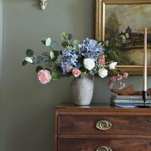 Pu&ograve; includere: Una composizione floreale in un vaso grigio &egrave; appoggiata su un com&ograve; in legno scuro. Il bouquet comprende ortensie blu, rose rosa e fiori bianchi. Un dipinto incorniciato e una candela sono anche sul com&ograve;.