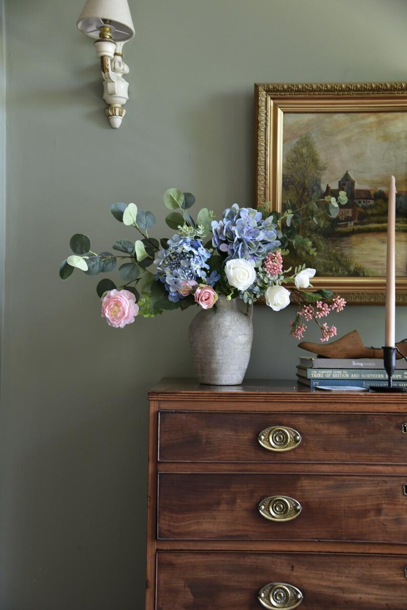 Pu&ograve; includere: Una composizione floreale in un vaso grigio &egrave; appoggiata su un com&ograve; in legno scuro. Il bouquet comprende ortensie blu, rose rosa e fiori bianchi. Un dipinto incorniciato e una candela sono anche sul com&ograve;.