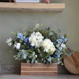 May include: A wooden box arrangement of white hydrangeas, blue flowers, and green foliage. The display is on a grey countertop. A framed landscape painting and a candle are on a wooden shelf in the background.