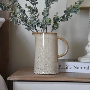 May include: A ceramic pitcher with a speckled glaze in shades of beige and brown, filled with a bouquet of green eucalyptus stems. The pitcher is sitting on a wooden nightstand with a white drawer and a gold knob. A book titled "Pacific Natural" is resting on the nightstand.