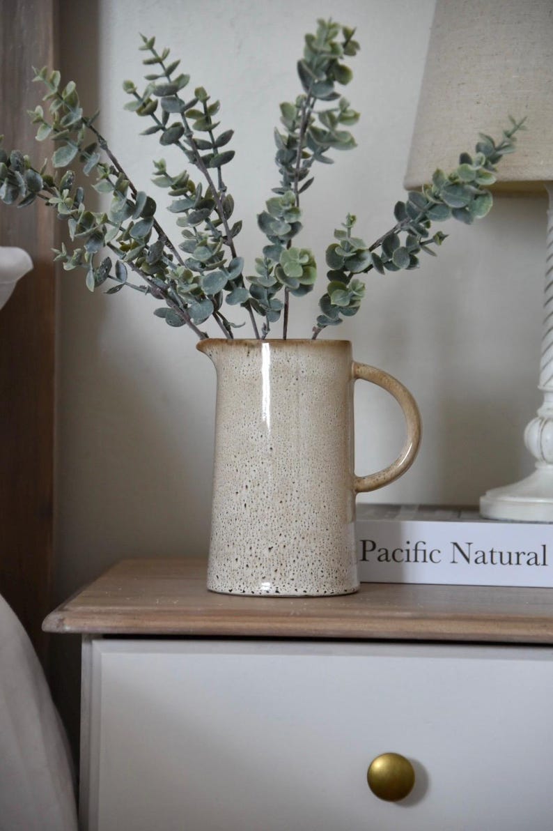 May include: A ceramic pitcher with a speckled glaze in shades of beige and brown, filled with a bouquet of green eucalyptus stems. The pitcher is sitting on a wooden nightstand with a white drawer and a gold knob. A book titled "Pacific Natural" is resting on the nightstand.