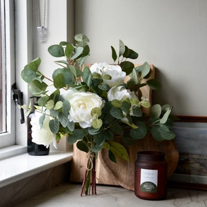 Puede incluir: Un ramo de peon&iacute;as blancas y hojas de eucalipto atadas con una cuerda, dispuestas sobre una tabla de cortar de madera con un tarro de vela de vidrio marr&oacute;n etiquetado como "The Est&eacute;e Candle Co. Eucalyptus & Amber Candle".