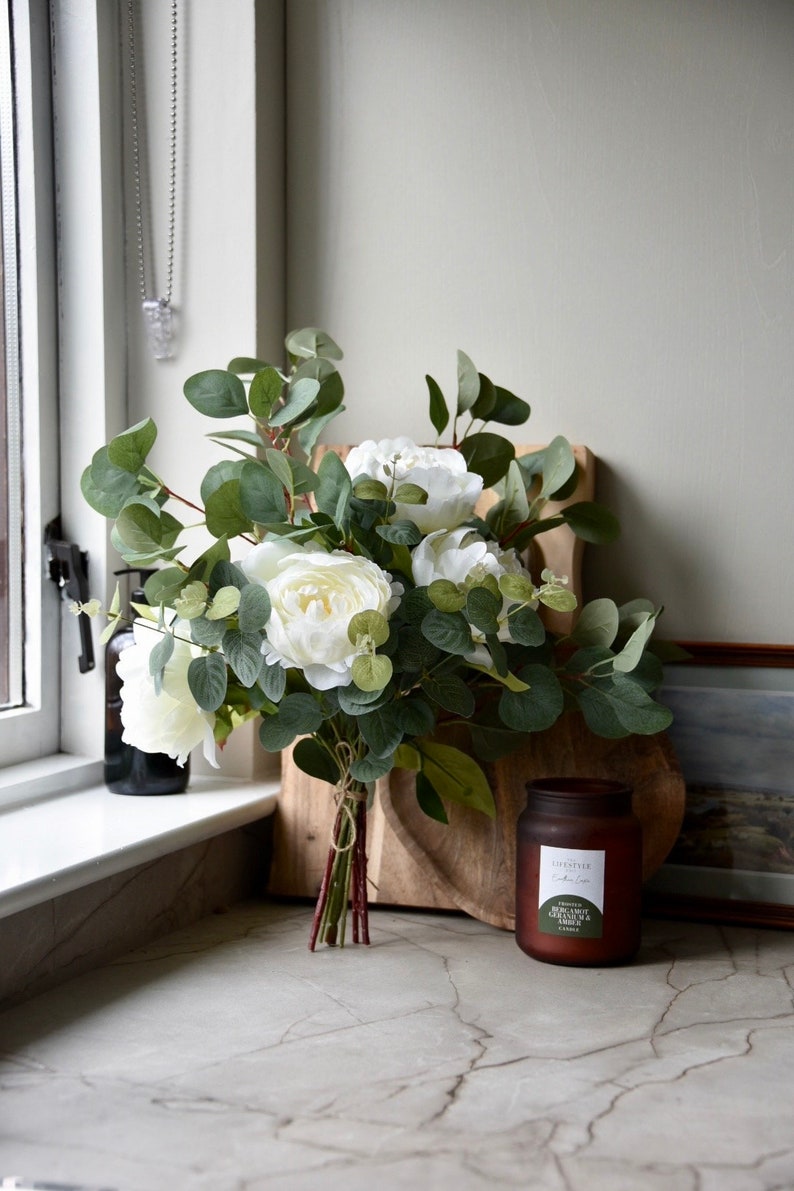 Puede incluir: Un ramo de peon&iacute;as blancas y hojas de eucalipto atadas con una cuerda, dispuestas sobre una tabla de cortar de madera con un tarro de vela de vidrio marr&oacute;n etiquetado como "The Est&eacute;e Candle Co. Eucalyptus & Amber Candle".