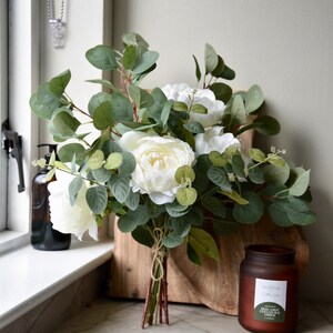 Puede incluir: Un ramo de peon&iacute;as blancas y hojas de eucalipto atadas con cuerda, sobre una tabla de cortar de madera junto a una vela de vidrio marr&oacute;n con una etiqueta verde que dice "A Lifestyle Collection - Herb Garden & Cedar Candle".