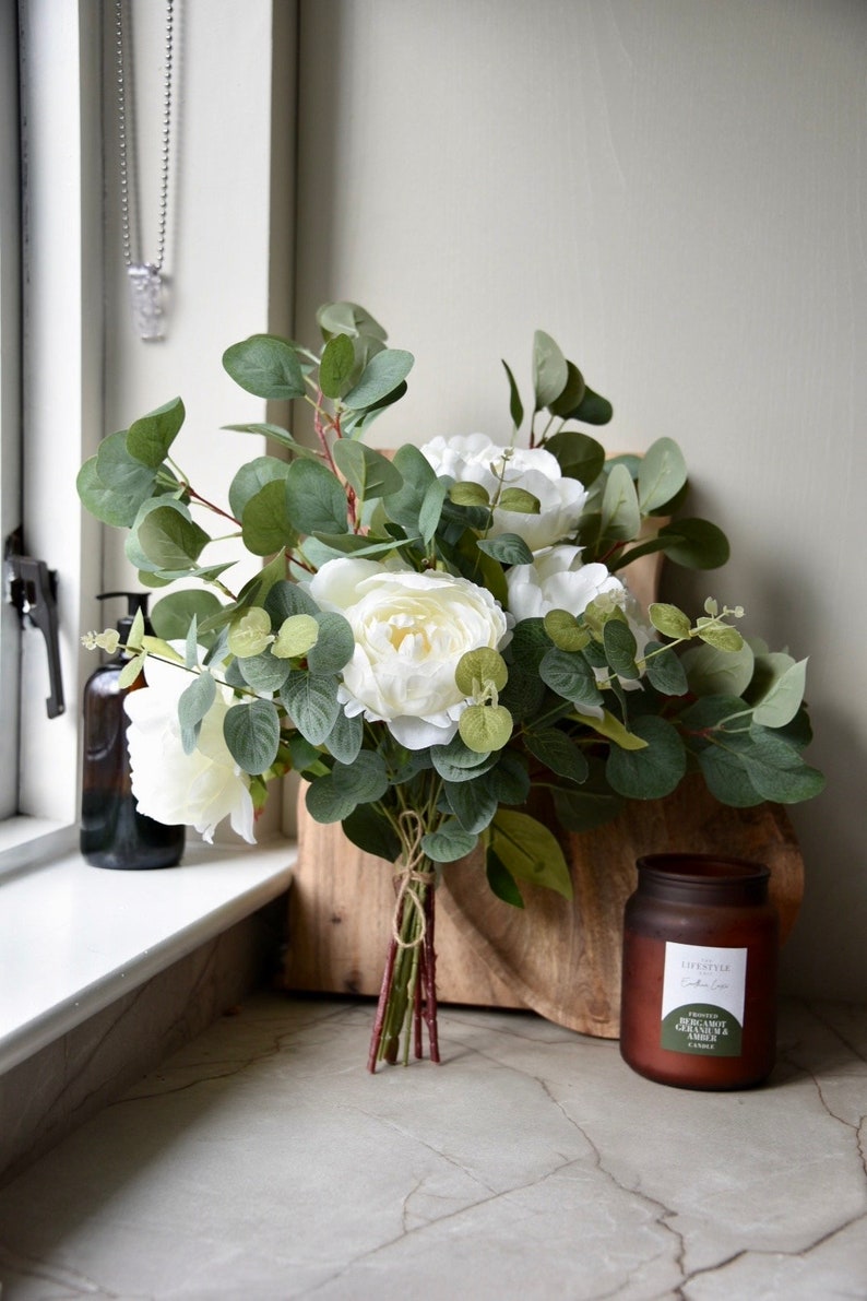 Puede incluir: Un ramo de peon&iacute;as blancas y hojas de eucalipto atadas con cuerda, sobre una tabla de cortar de madera junto a una vela de vidrio marr&oacute;n con una etiqueta verde que dice "A Lifestyle Collection - Herb Garden & Cedar Candle".