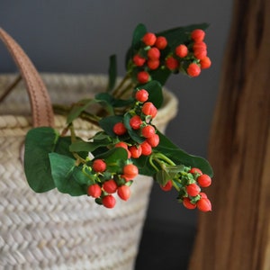 Może przedstawiać: Kosz wiklinowy z brązowym skórzanym uchwytem wypełniony gałązką czerwonych jagód i zielonych liści.
