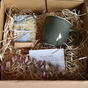 May include: A gift box with a green ceramic mug, a small framed photograph of a path, and a card with the text "Wool Hill FINE ART & HOMEWARE". The box is filled with straw and decorative sprigs.