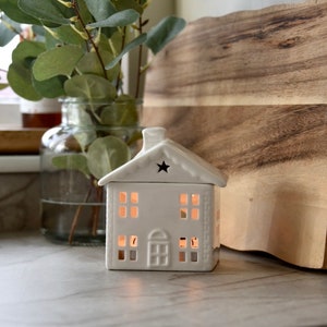 Puede incluir: Un portavelas de cerámica blanca con forma de casa con chimenea y un recorte de estrella. La casa tiene múltiples recortes de ventanas que brillan con la luz de las velas en su interior. Un jarrón de cristal con hojas verdes y una tabla de cortar de madera están en el fondo.