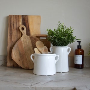 May include: A kitchen countertop arrangement with wooden chopping boards, white ceramic pots, wooden spoons, and a vase with greenery. A brown bottle with a pump dispenser is also present. The items are on a marble-effect surface.