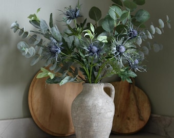 Puede incluir: Un arreglo floral en un jarrón gris claro texturizado. El ramo presenta flores azules parecidas al cardo y hojas verdes de eucalipto. Dos tablas de cortar de madera están en el fondo, creando una estética rústica y natural.