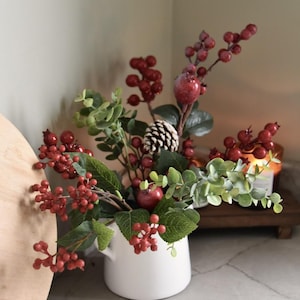 Könnte beinhalten: Eine weiße Keramikkrug mit einem herbstlichen Arrangement. Das Arrangement umfasst rote Beeren, grüne Eukalyptusblätter, kleine rote Äpfel und einen Tannenzapfen. Der Hintergrund ist neutral gefärbt.