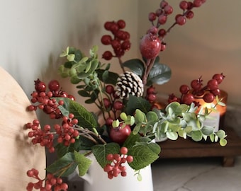 May include: A white ceramic pitcher filled with a fall-themed arrangement. The arrangement includes red berries, green eucalyptus leaves, small red apples, and a pinecone. The background is a neutral color.