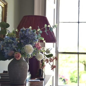 Pu&ograve; includere: Una composizione floreale in un vaso grigio con ortensie blu, rose bianche e accenti rosa. Un paralume bordeaux &egrave; dietro il vaso su una superficie in legno scuro. Sullo sfondo una finestra con vista sul verde.